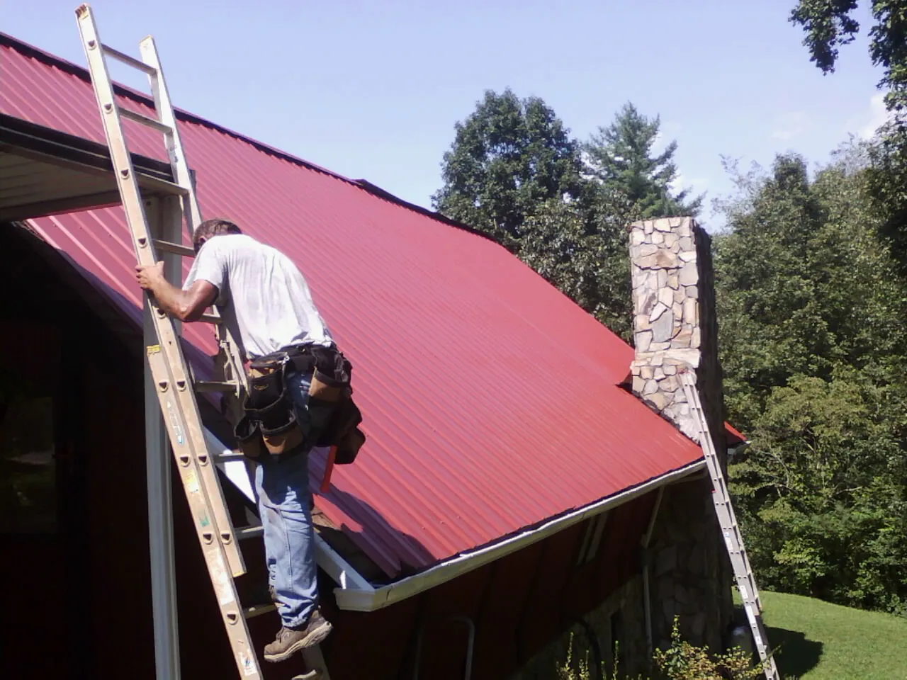 Roofer on a ladder beside a red metal roof and stone chimney — Soffit & Fascia Repair by Roofing Brothers in Dodgeville
